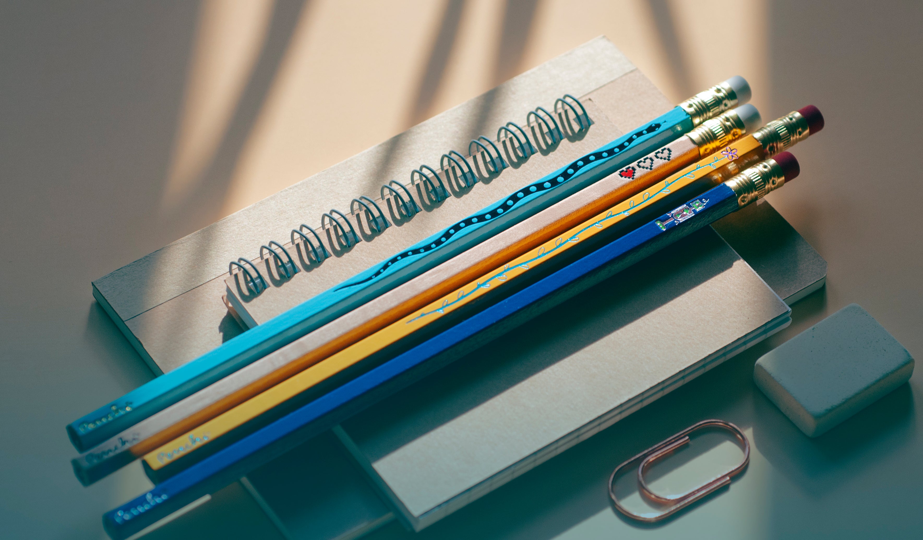 Penchi hand-stamped metallic foil pencils featuring creative visual designs like snakes, hearts, flowers and robots, paired with other stationery items like a notebook, eraser, and paper clip